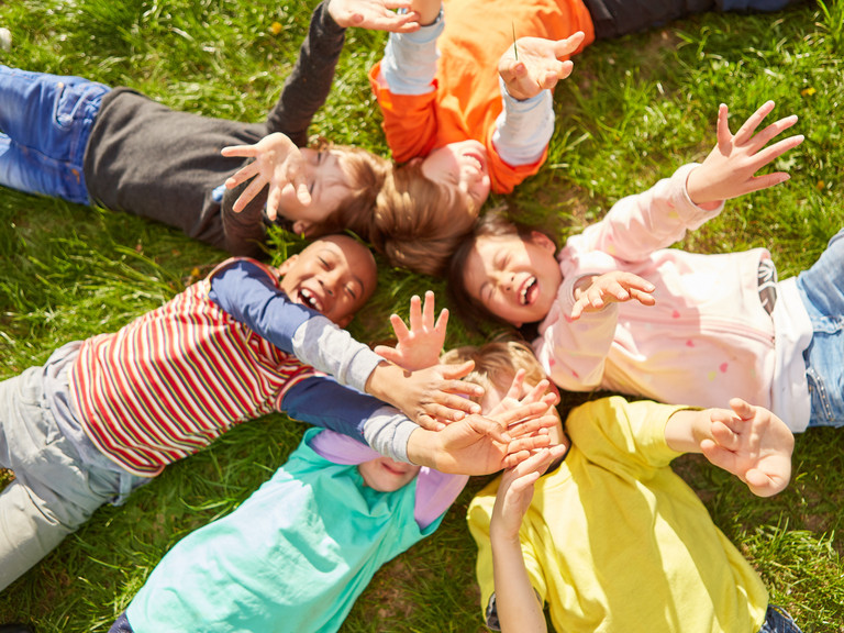 Sechs Kinder liegen lachend im Kreis auf einer grünen Wiese und strecken ausgelassen ihre Arme in die Luft.
