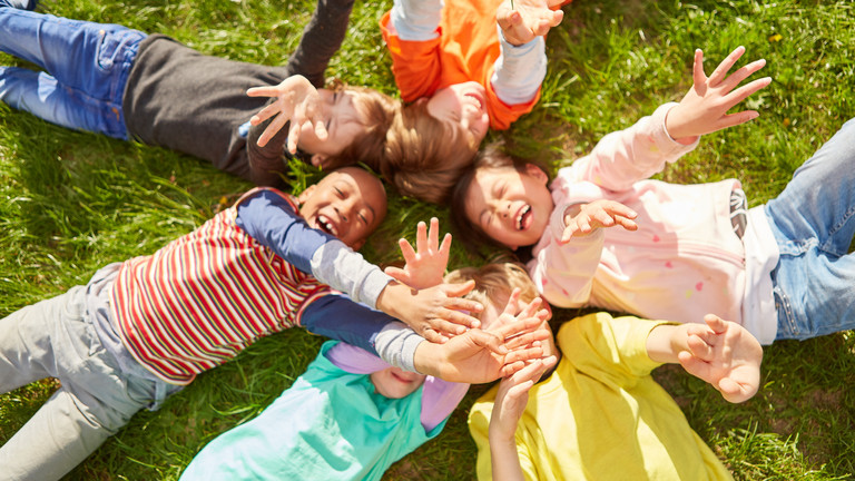 Sechs Kinder liegen lachend im Kreis auf einer grünen Wiese und strecken ausgelassen ihre Arme in die Luft.