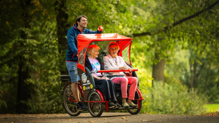 Ein Fahrer fährt zwei Seniorinnen mit einer roten E-Rikscha von fLotte SoziaL durch einen grünen Park mit Bäumen und Blättern im Berliner Bezirk Tempelhof-Schöneberg mit dem E-Rikscha-Standort beim Immanuel Stadtteilzentrum Schöneberg.