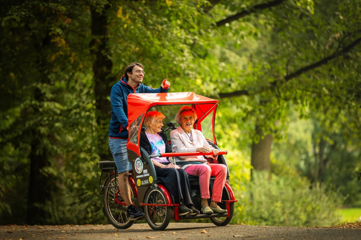 Ein Fahrer fährt zwei Seniorinnen mit einer roten E-Rikscha von fLotte SoziaL durch einen grünen Park mit Bäumen und Blättern im Berliner Bezirk Tempelhof-Schöneberg mit dem E-Rikscha-Standort beim Immanuel Stadtteilzentrum Schöneberg.