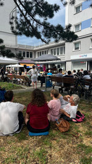 Zahlreiche Menschen sitzen auf einer Sitzwiese beim Musikfestival Fête de la Musique um den Platz vor dem Immanuel Stadtteilzentrum Borsigwalde in Berlin-Reinickendorf.