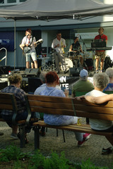Zuschauer sitzen auf Sitzbänken vor einer Musikbühne mit einer Band beim Musikfestival Fête de la Musique im Immanuel Stadtteilzentrum Borsigwalde in Berlin-Reinickendorf.