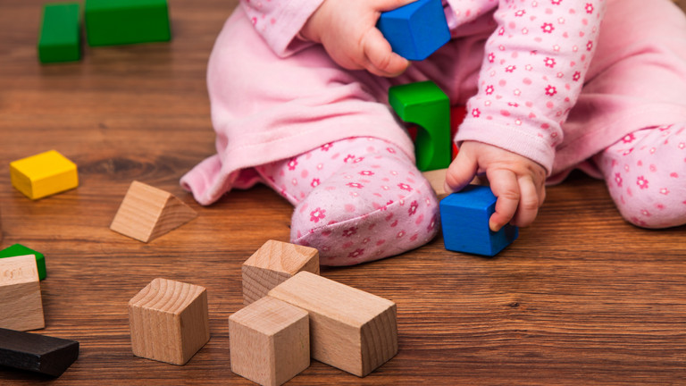 Ein Kleinkind sitzt mit einem hellpinken Overall auf einem Holzboden und spielt mit bunten Holzklötzen als Motiv für den Kurs für Eltern mit Baby FuN-Baby der Immanuel Beratung Fennpfuhl mit dem Kinder-und Jugendgesundheitsdienst Lichtenberg (KJGD) in Berlin. Ein Kleinkind sitzt mit einem hellpinken Overall auf einem Holzboden und spielt mit bunten Holzklötzen als Motiv für den Kurs für Eltern mit Baby FuN-Baby der Immanuel Beratung Fennpfuhl mit dem Kinder-und Jugendgesundheitsdienst Lichtenberg (KJGD) in Berlin.