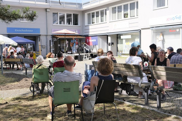 Mehrere Menschen sitzen auf Klappstühlen und Sitzbänken um den Platz mit einer Musikbühne beim Musikfestival Fête de la Musique vor dem Immanuel Stadtteilzentrum Borsigwalde in Berlin-Reinickendorf.
