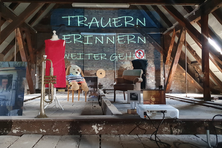 Ein Dachboden mit mehreren Gegenständen, ein Beamer wirft die Wörter "Trauern, Erinnern, Weitergehen" an die Wand