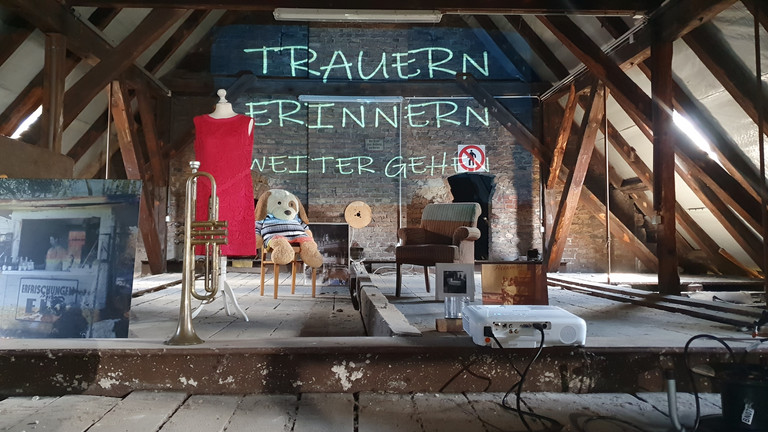 Ein Dachboden mit mehreren Gegenständen, ein Beamer wirft die Wörter "Trauern, Erinnern, Weitergehen" an die Wand